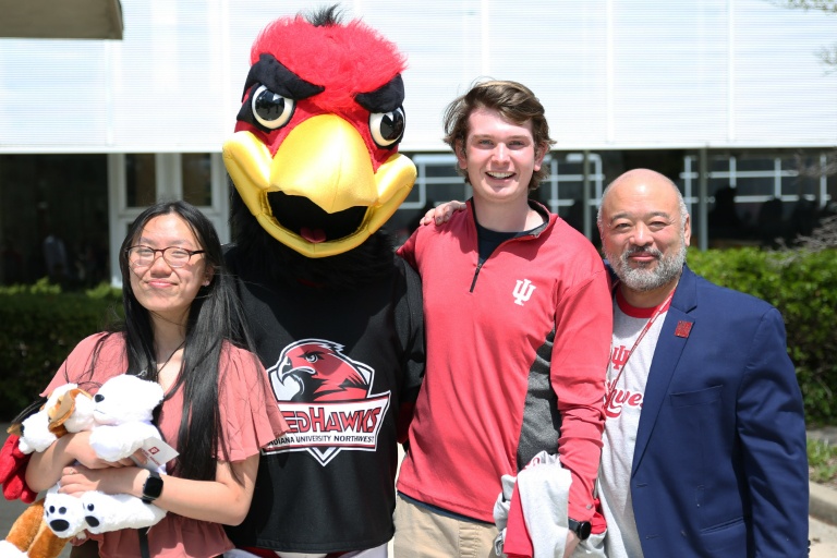 Four, one dressed in a red bird costume, stand smiling outdoors.